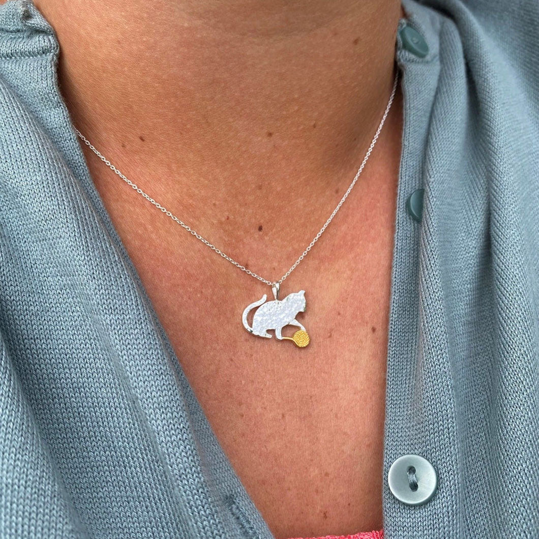 Necklace with a cat and tennis ball pendant worn by a person in a light blue shirt.