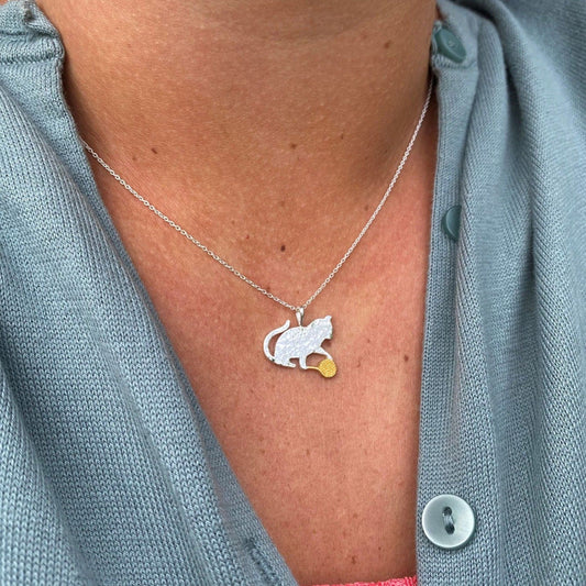 Necklace with a cat and tennis ball pendant worn by a person in a light blue shirt.