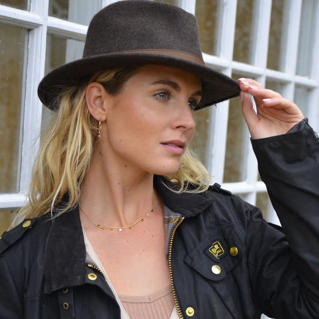 Woman wearing a black hat and black jacket with gold buttons, standing in front of a window.