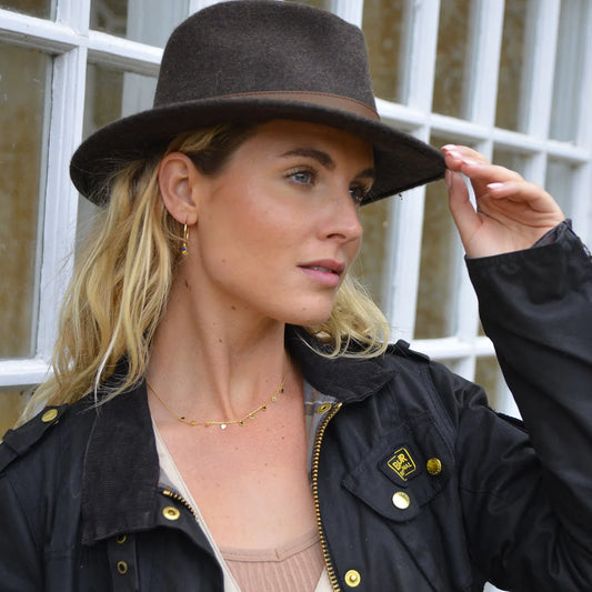 Woman wearing a black hat and black jacket with gold buttons, standing in front of a window.