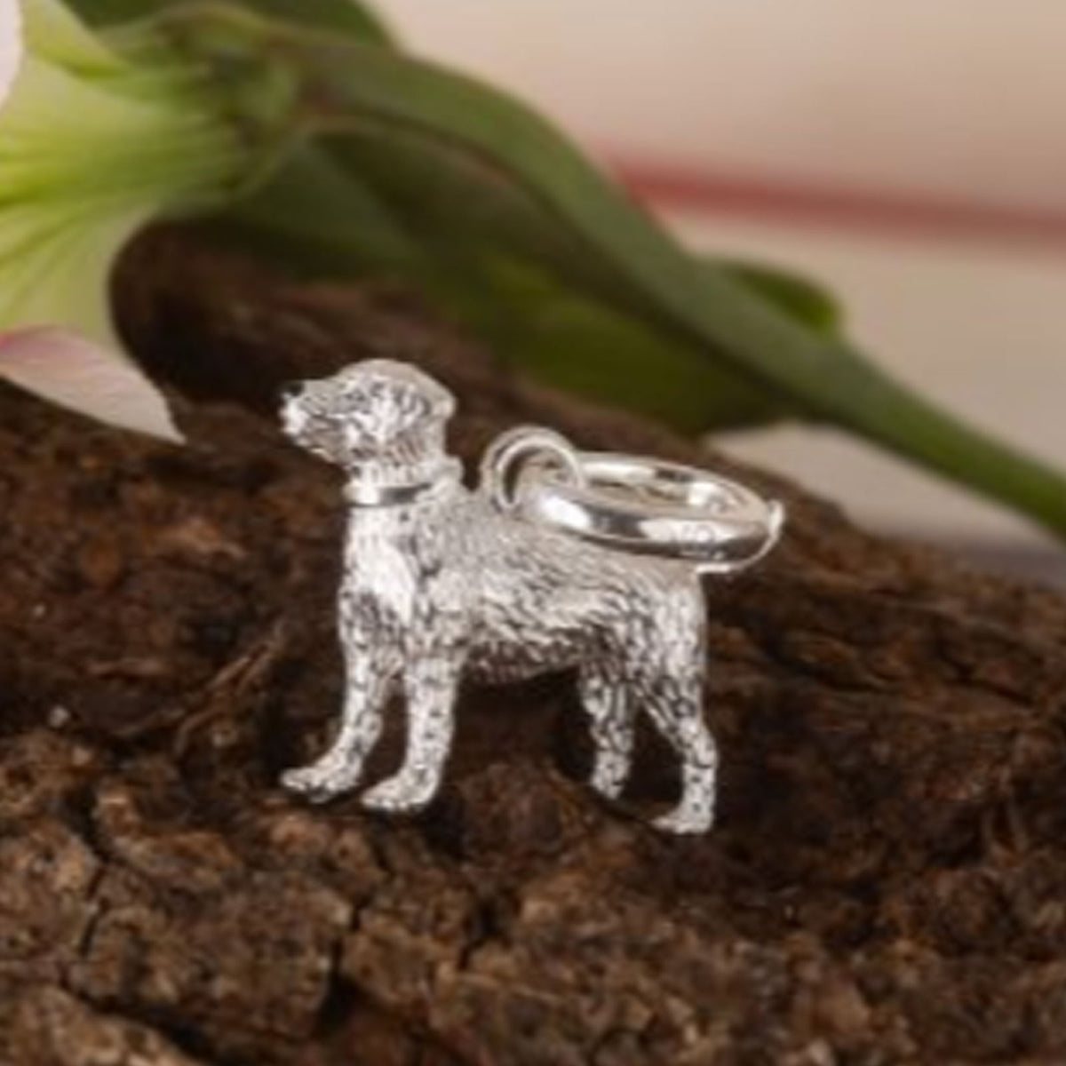 A silver dog-shaped charm or pendant with a textured finish and a smooth ring loop, displayed on a piece of bark with blurred green leaves in the background.