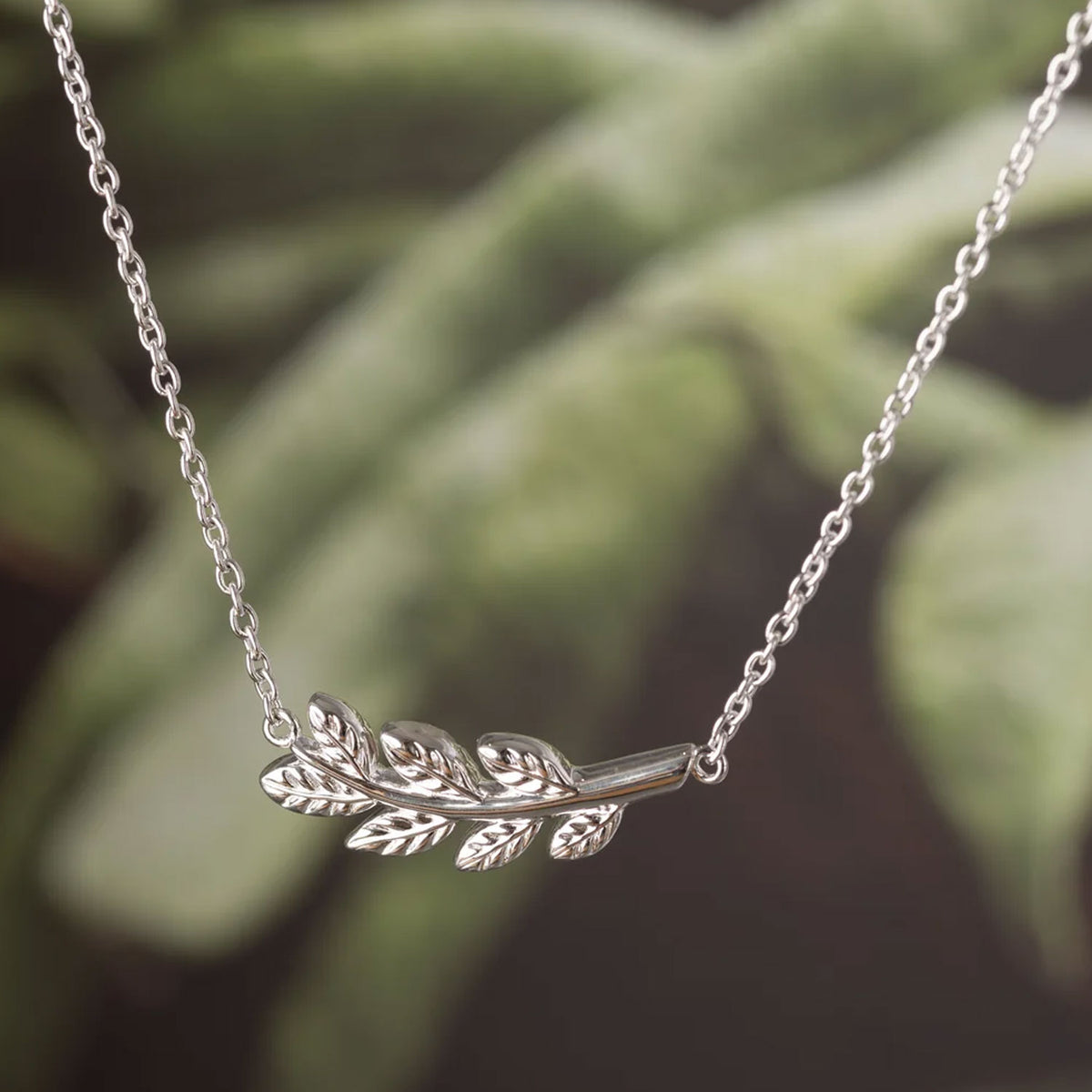A silver laurel leaf branch pendant necklace on a delicate silver chain, shown against a soft, blurred green background.