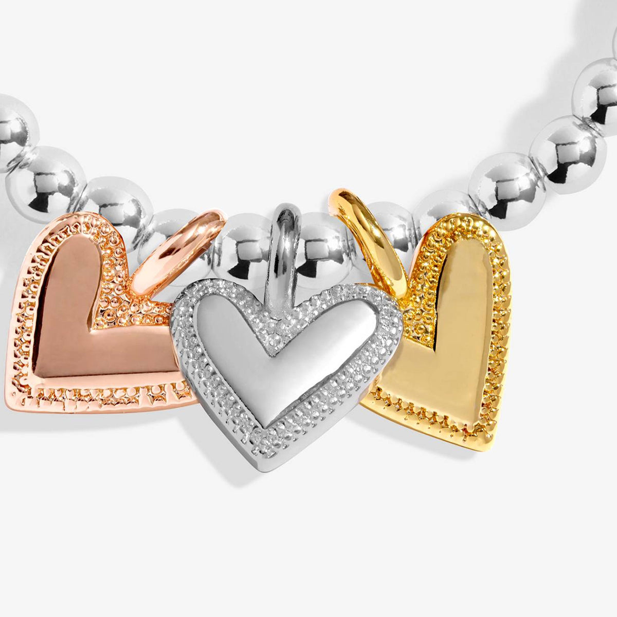 A close-up of a beaded bracelet featuring three heart charms in rose gold, yellow gold, and silver, with the center silver heart encrusted with small clear stones.