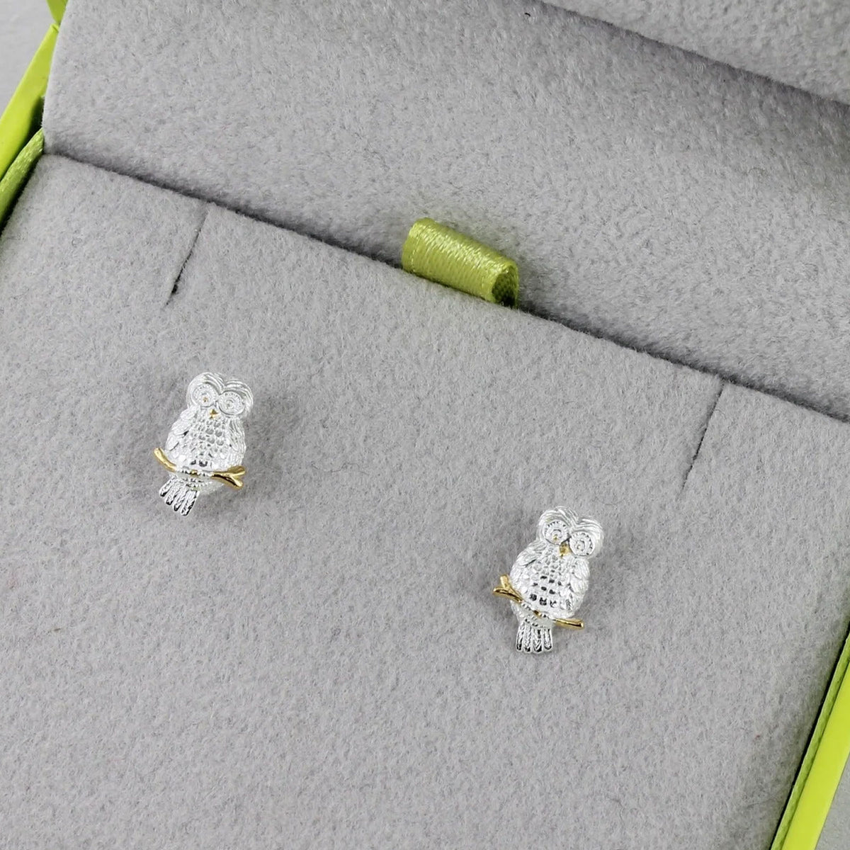 A pair of silver owl stud earrings with clear crystal-like detailing and small gold accents, displayed on a gray jewelry box insert.