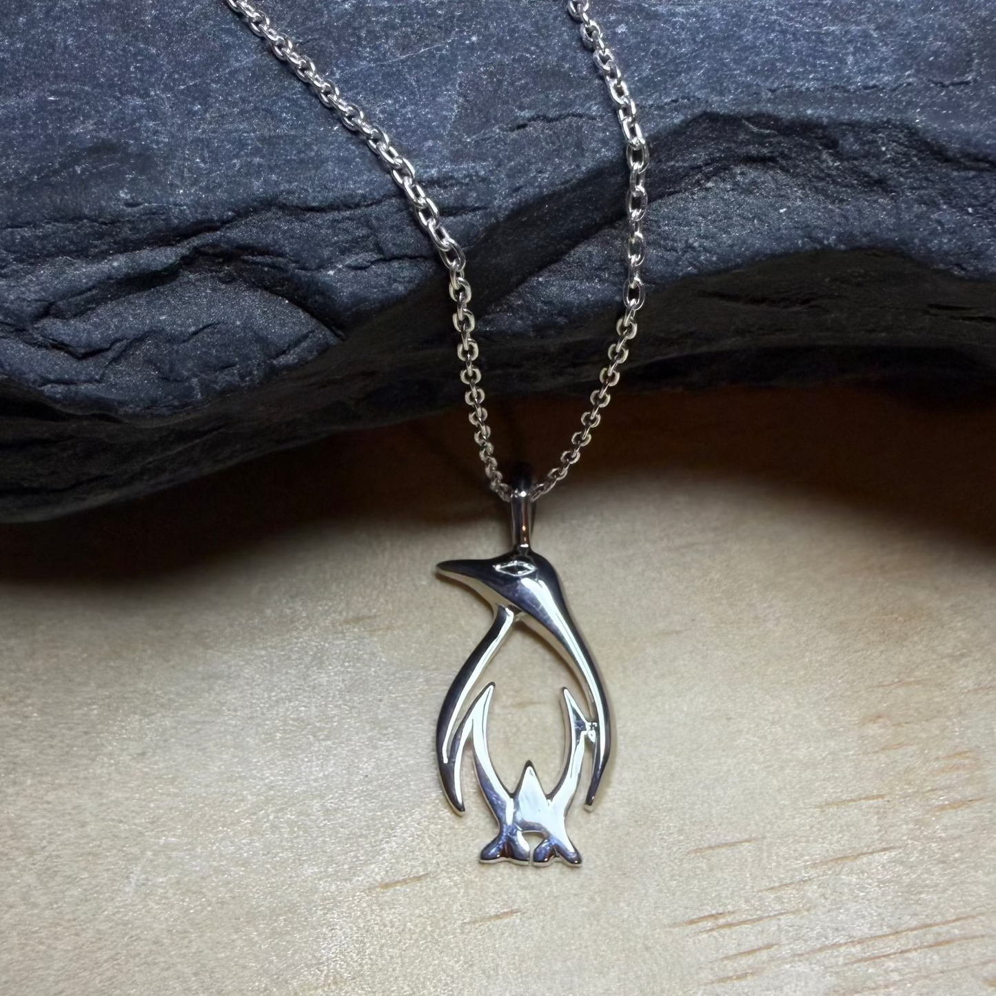 Silver penguin pendant on a chain against a stone and wooden background
