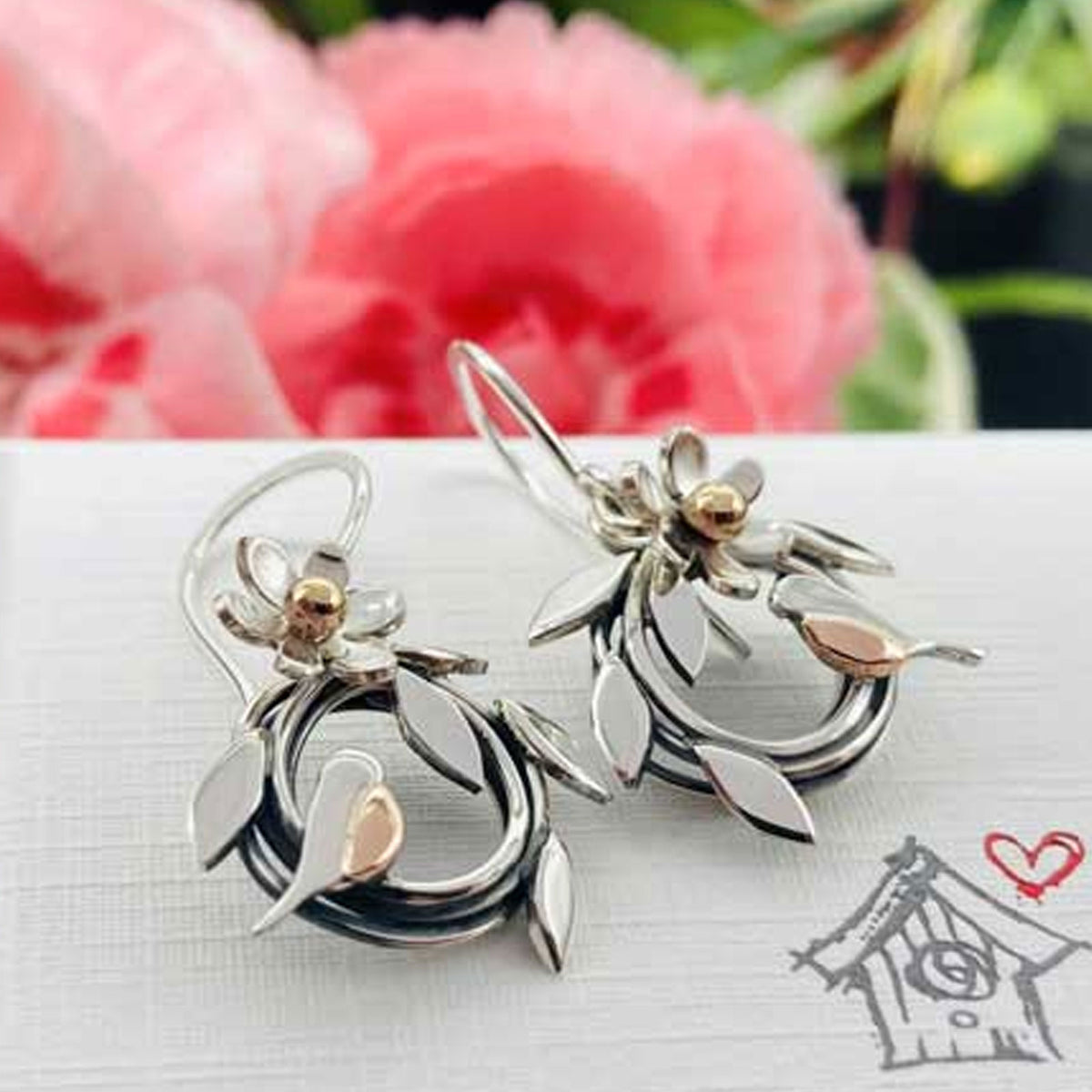 A pair of silver-toned dangle earrings with floral and leaf motifs, featuring small rose gold accents, displayed on a white textured surface with a blurred pink flower in the background.