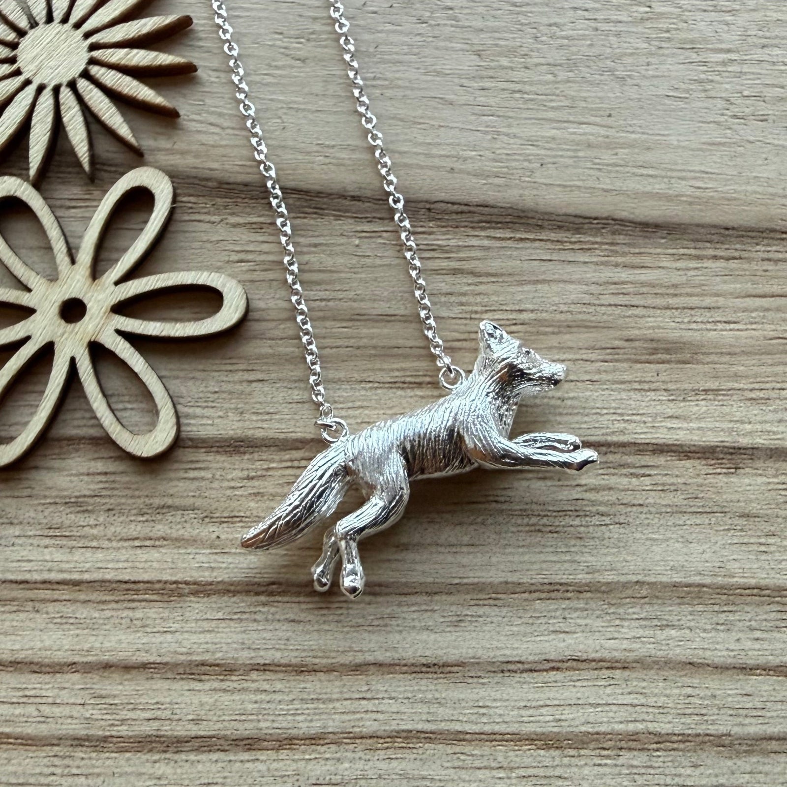 A silver pendant necklace featuring a detailed running fox on a delicate silver chain, displayed on a light wood grain background with decorative wooden flower cutouts.