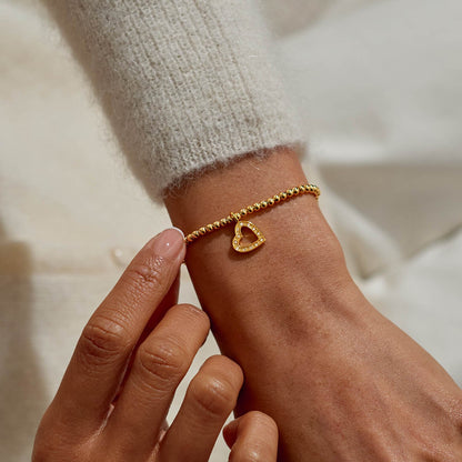 Gold bracelet with a heart charm on a wrist against a neutral background