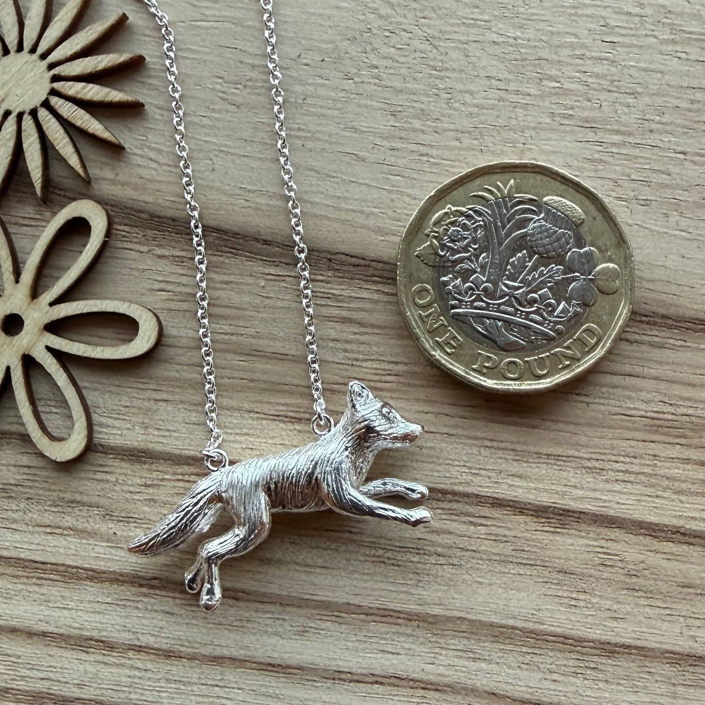A sterling silver fox pendant necklace on a delicate chain, displayed on a light wood surface next to a British penny for scale and wooden flower cutouts.