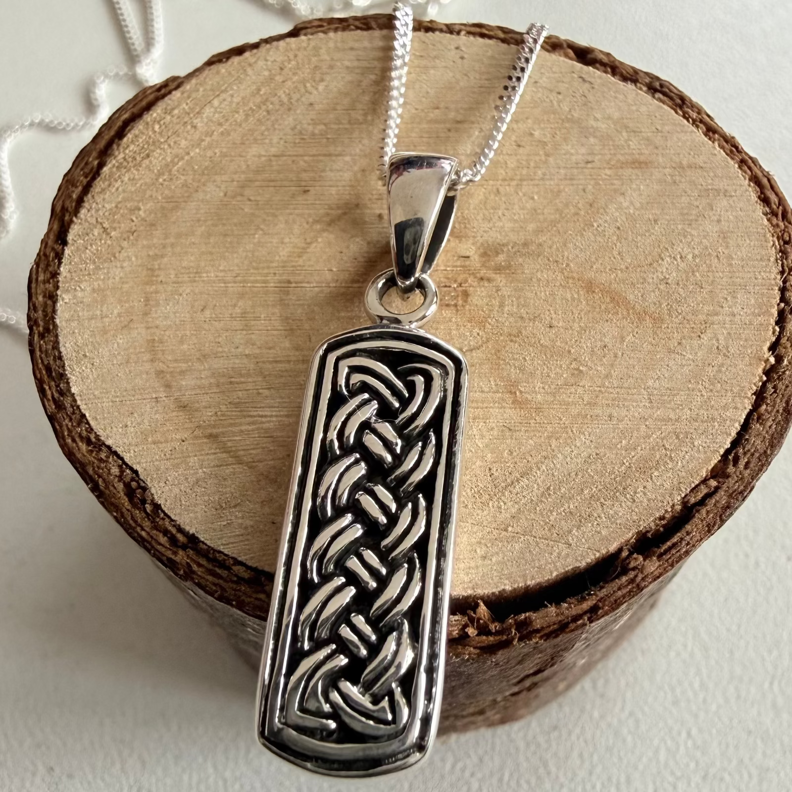 A silver pendant necklace with an engraved Celtic knot design, displayed on a rustic wood slice stand against a light gray background.
