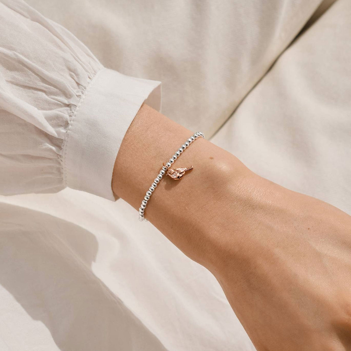 A delicate bracelet worn on a person's wrist, featuring a small rose gold bird charm on a silver beaded chain. The person is wearing a white long-sleeve blouse against a soft, light-colored fabric background.