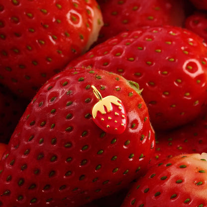 A small strawberry-shaped charm with a gold loop at the top, resting on a glossy red strawberry among more strawberries.