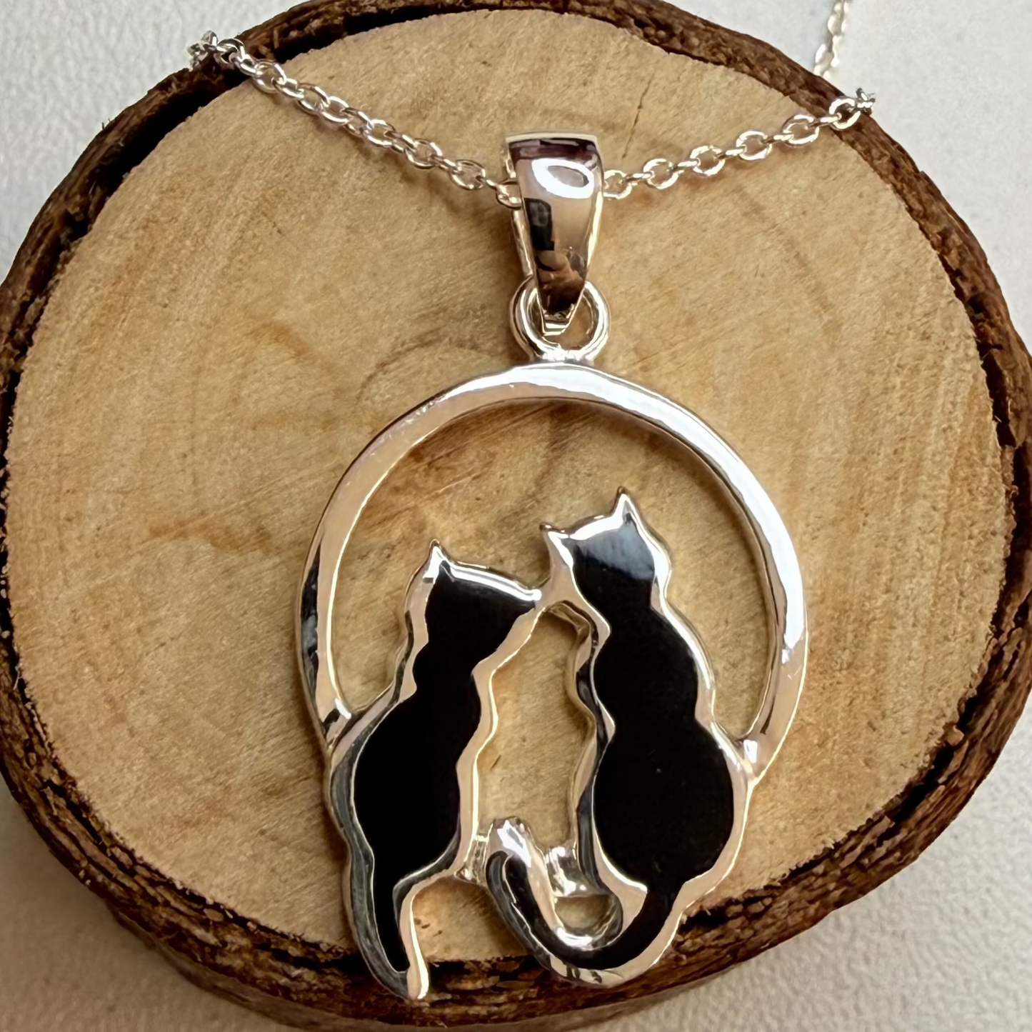 A silver pendant necklace featuring two black cat silhouettes inside a circular frame, displayed on a round wood slice against a light beige background.