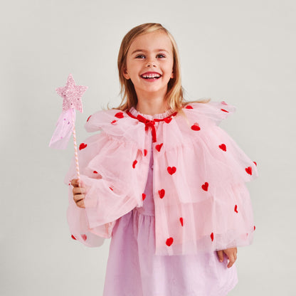 Young girl in a pink dress with red heart patterns, holding a star-shaped wand against a plain background.
