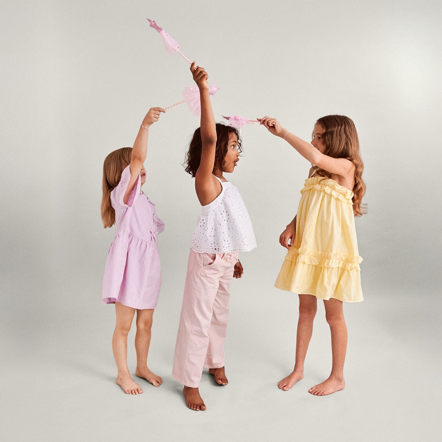 Three young girls in pastel dresses and pants holding pink fairy wands against a light background