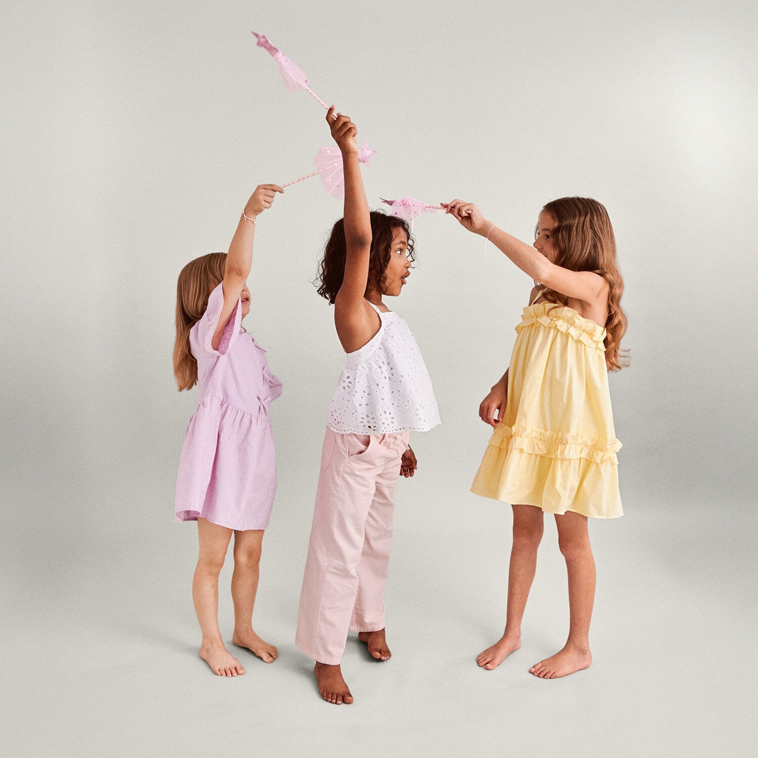 Three young girls in pastel dresses and pants holding pink fairy wands against a light background