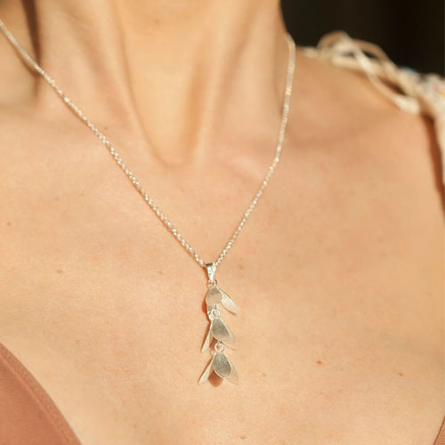 A close-up of a delicate silver leaf pendant necklace worn on a model's neck and chest, featuring three layered textured leaves on a fine silver chain.