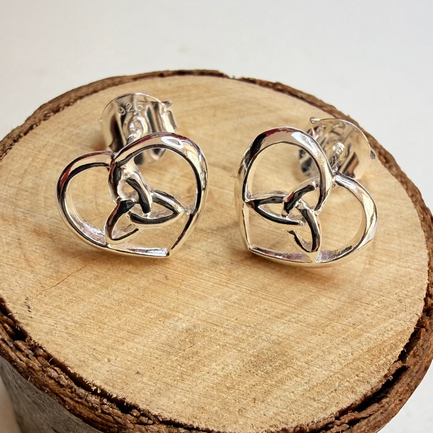 A pair of silver heart-shaped triquetra knot stud earrings displayed on a rustic wood slice prop against a light off-white background.