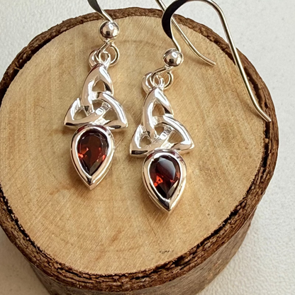 A pair of silver Celtic knot dangle earrings with teardrop-shaped dark red gemstones, displayed on a rustic wooden slice against a light background.