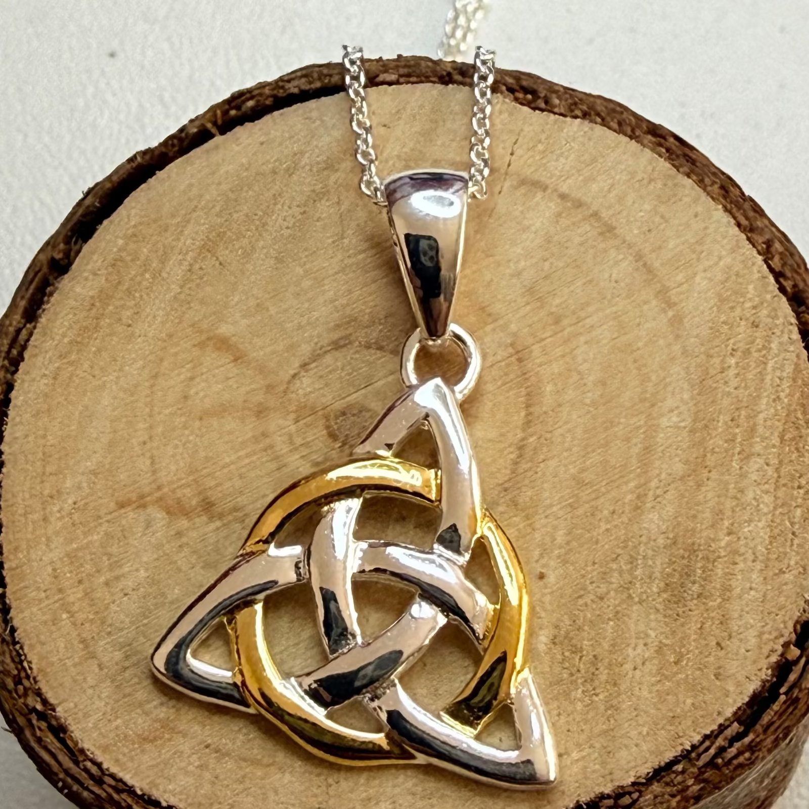 A two-tone silver and gold Celtic triquetra pendant necklace displayed on a rustic wood slice background.