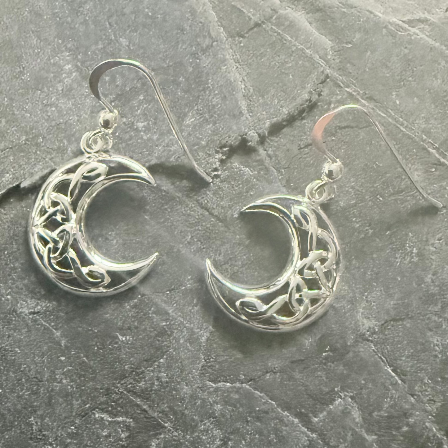 A pair of silver crescent moon dangle earrings with Celtic knotwork designs, displayed on a textured gray slate surface.