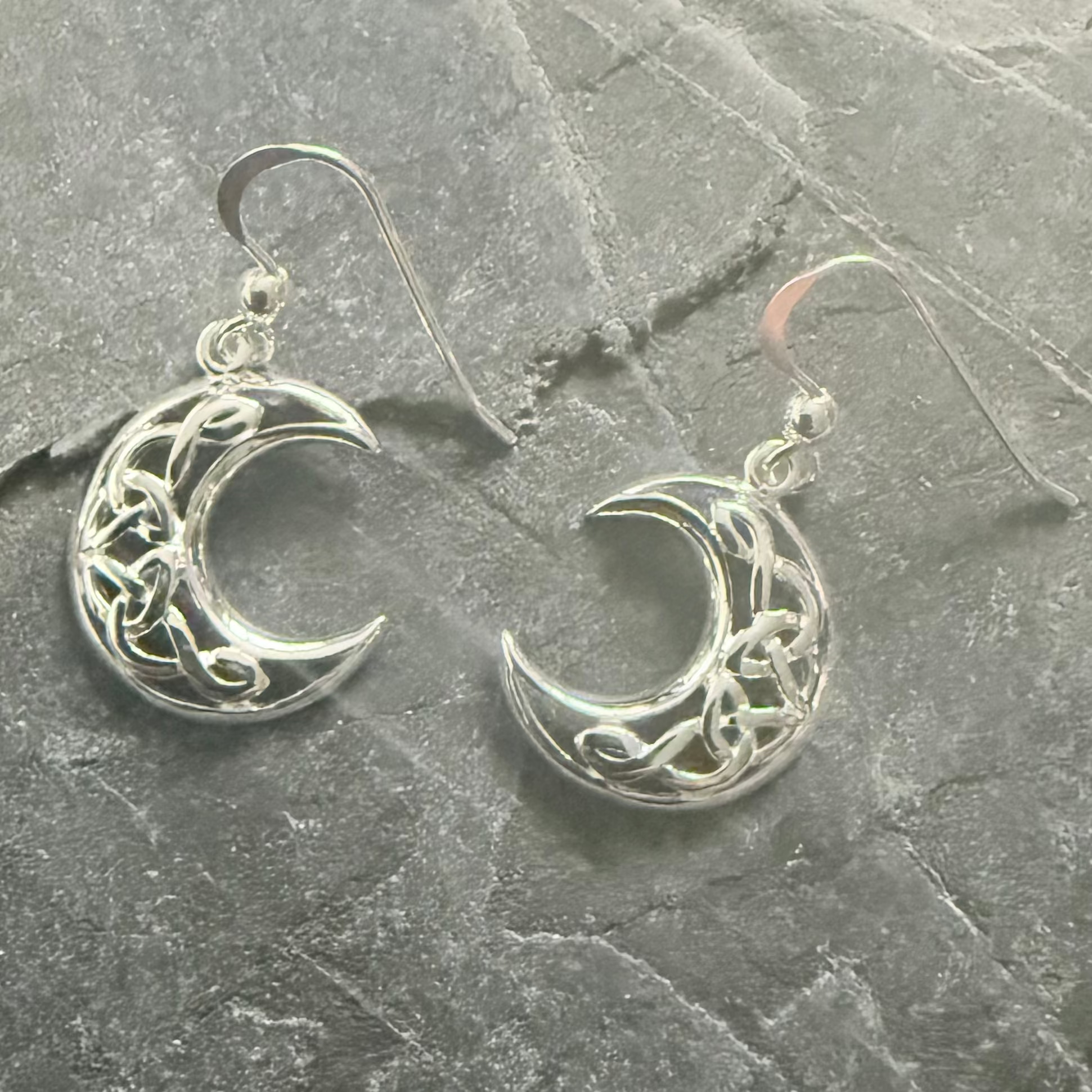 A pair of silver crescent moon dangle earrings with Celtic knotwork designs, displayed on a textured gray slate surface.