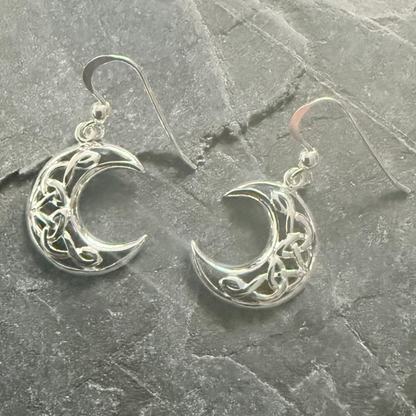 A pair of silver crescent moon dangle earrings with Celtic knotwork designs, displayed on a textured gray slate surface.