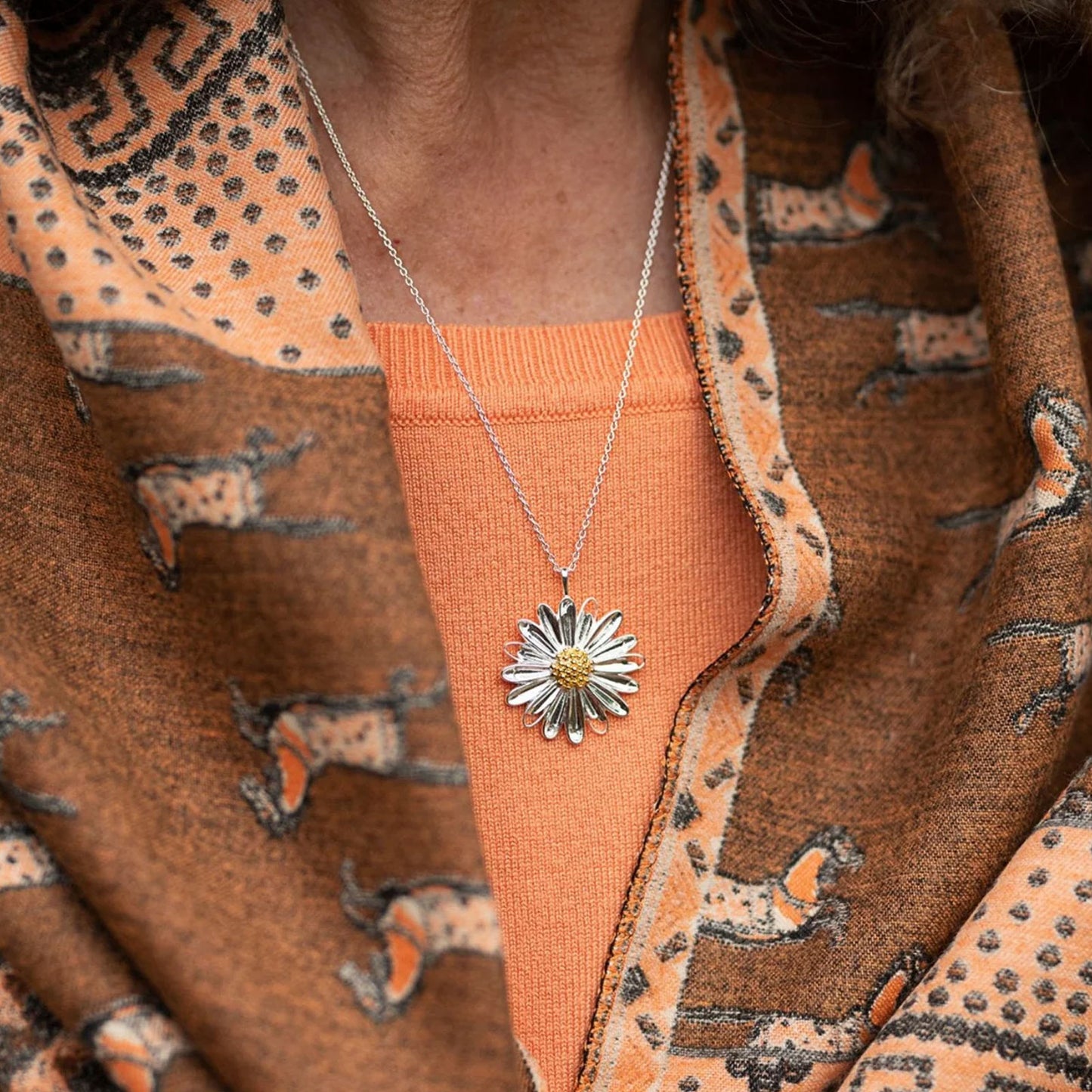 A close-up of a person wearing a silver daisy pendant necklace over an orange top, layered under a brown and orange patterned knit cardigan.
