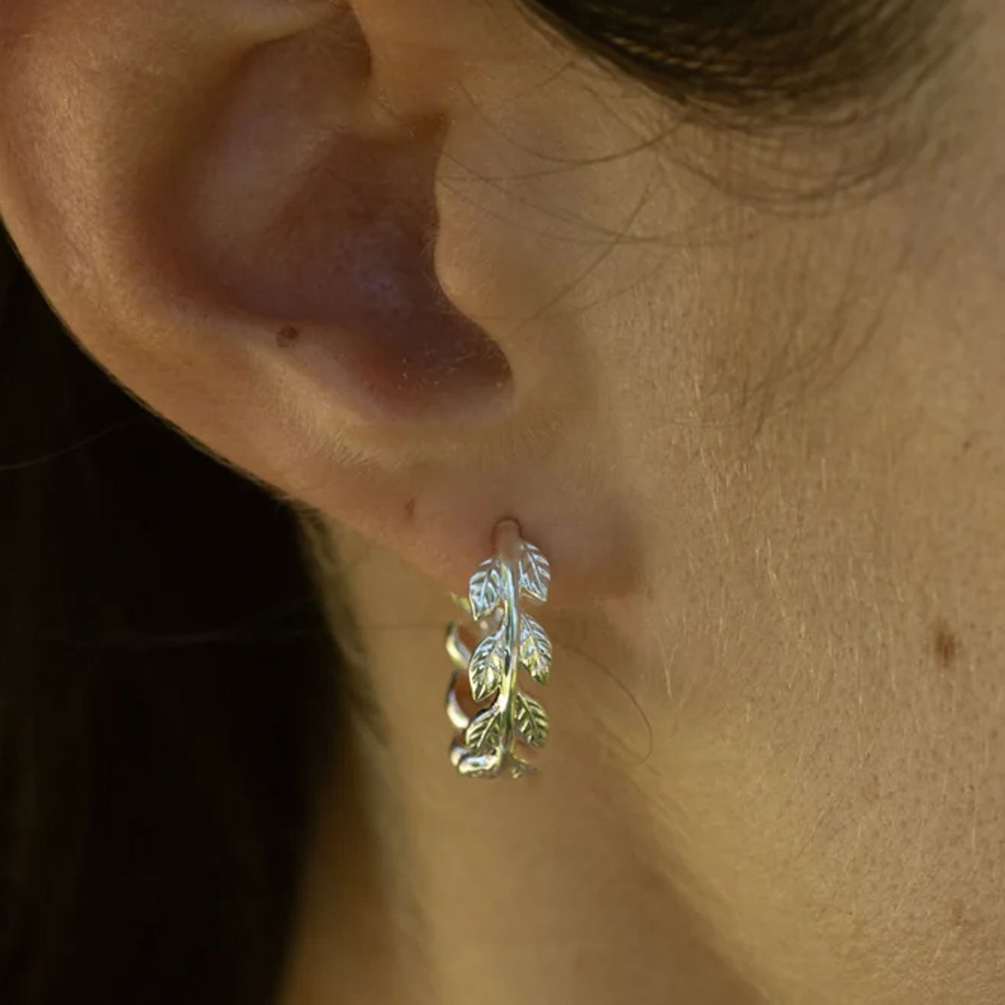 A close-up of a person's ear wearing a small silver huggie hoop earring with a leaf design.