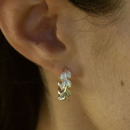 A close-up of a person's ear wearing a small silver huggie hoop earring with a leaf design.