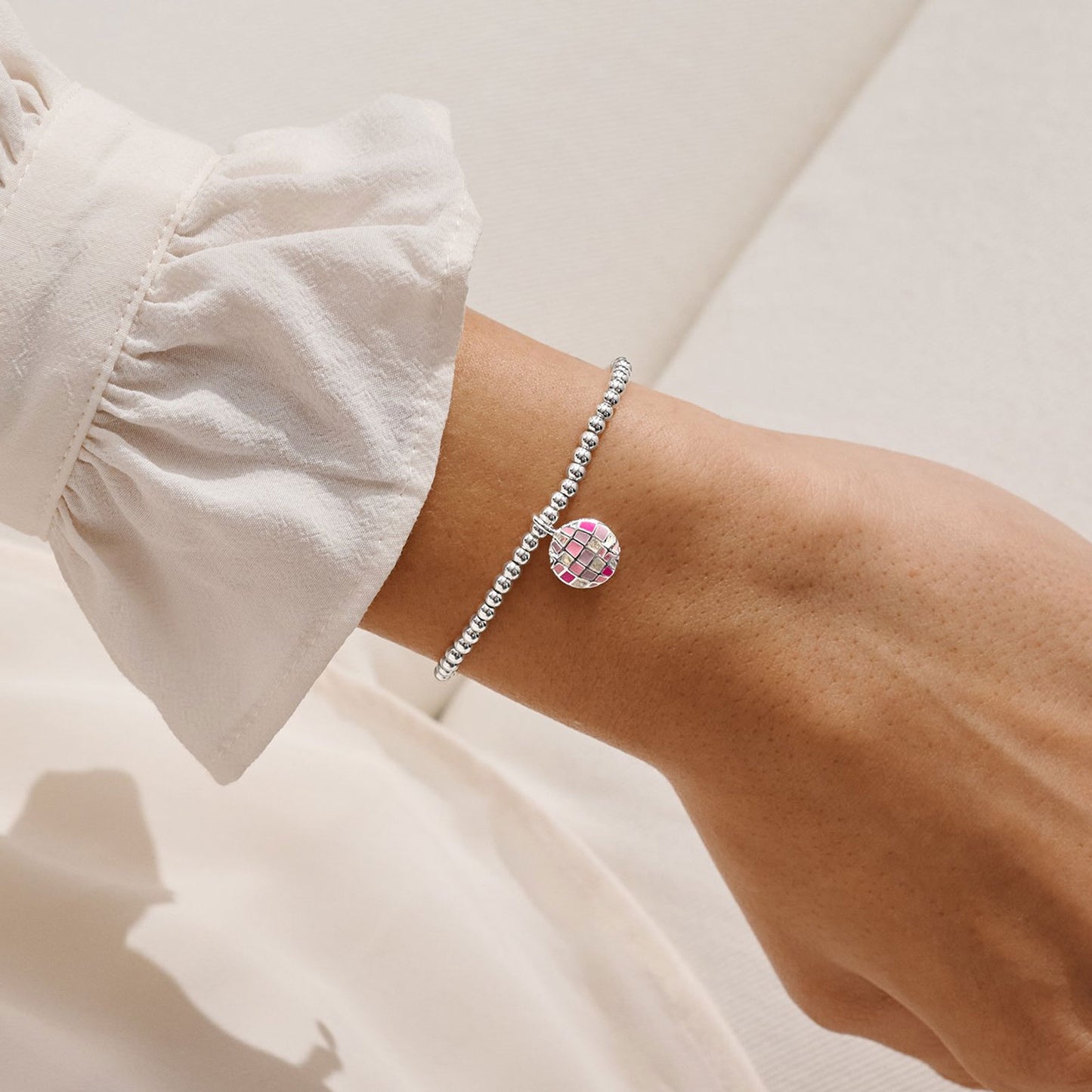 A close-up of a person's wrist wearing a delicate silver beaded bracelet with a round pink and green floral charm, styled with a white ruffled sleeve.