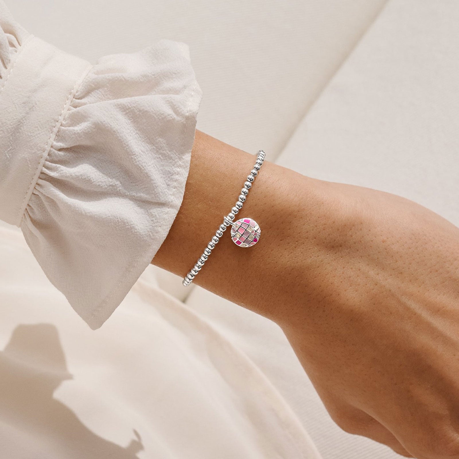 A close-up of a person's wrist wearing a delicate silver beaded bracelet with a round pink and green floral charm, styled with a white ruffled sleeve.