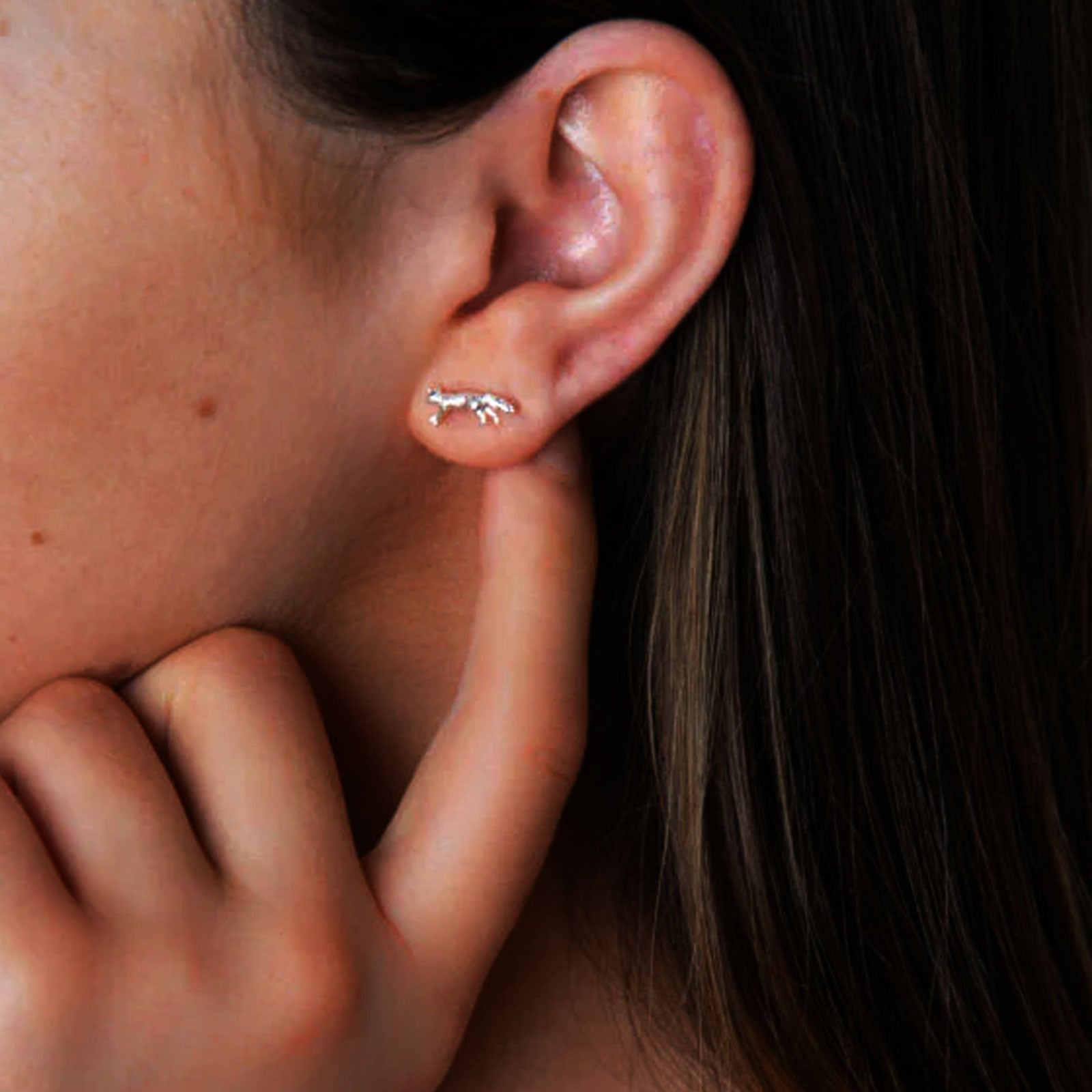 Close-up of a person's ear with a small fox earring, hand touching the earlobe.