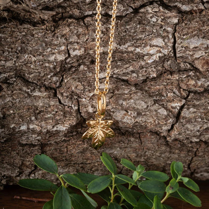 Gold necklace with a leaf-shaped pendant on a wooden surface with green leaves and a tree trunk background.