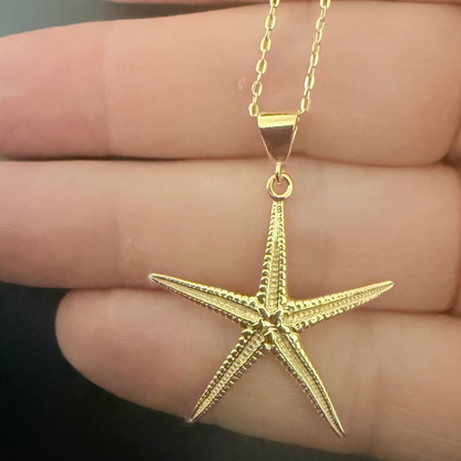 A gold starfish pendant necklace held in a person's hand against a dark background.