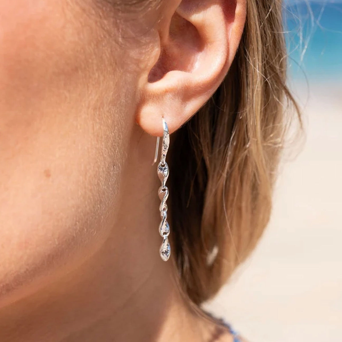 A close-up of a person's ear wearing a silver drop wave inspired earring on a blurred beach background.