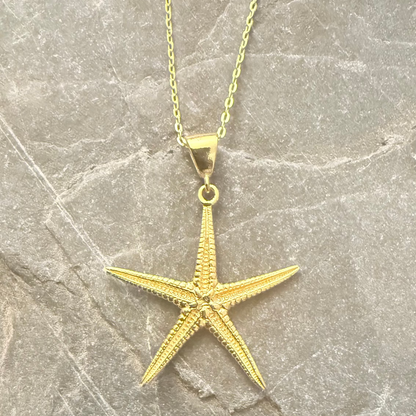 A gold starfish pendant necklace on a matching chain, displayed on a textured gray stone surface.