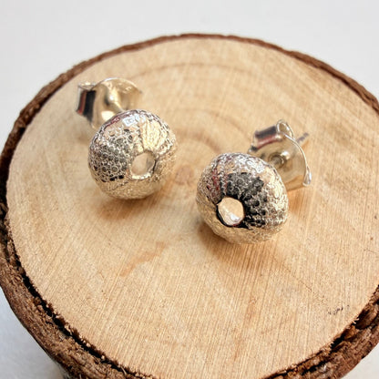 A pair of textured silver sea urchin stud earrings displayed on a round wood slice coaster against a light off-white background.