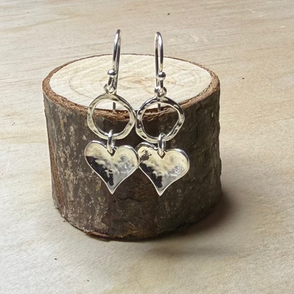 Silver heart-shaped earrings on a wooden block with a desk and supplies in the background.