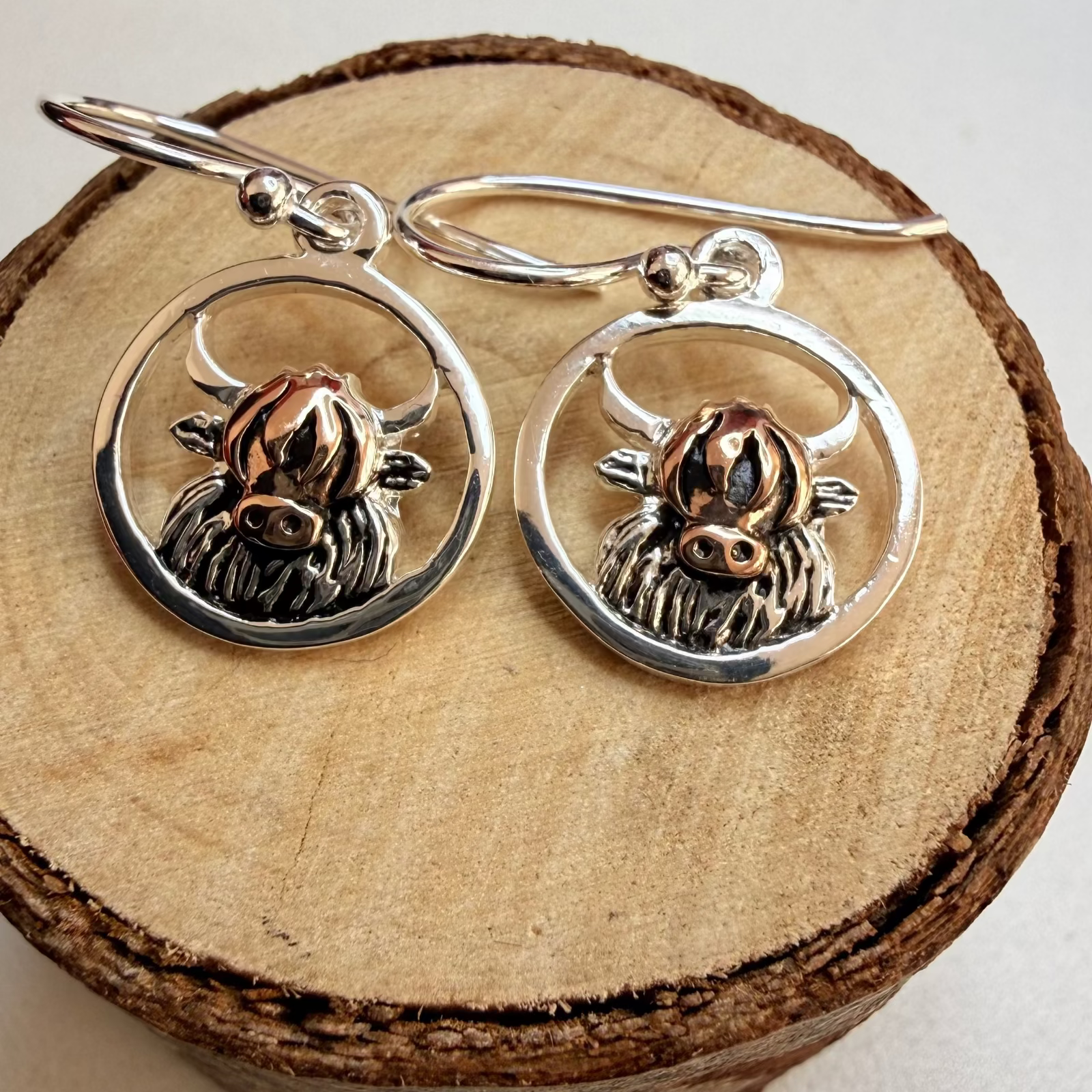 A pair of sterling silver dangle earrings with circular frames and detailed  Highland cow charms, displayed on a rustic wood slice against a light neutral background.