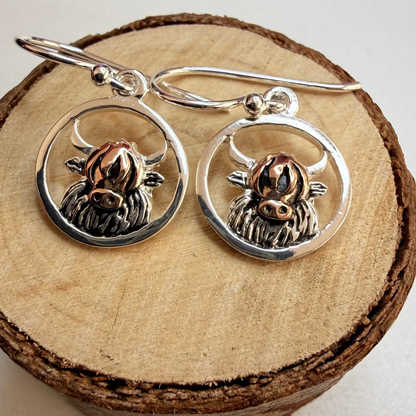A pair of sterling silver dangle earrings with circular frames and detailed  Highland cow charms, displayed on a rustic wood slice against a light neutral background.