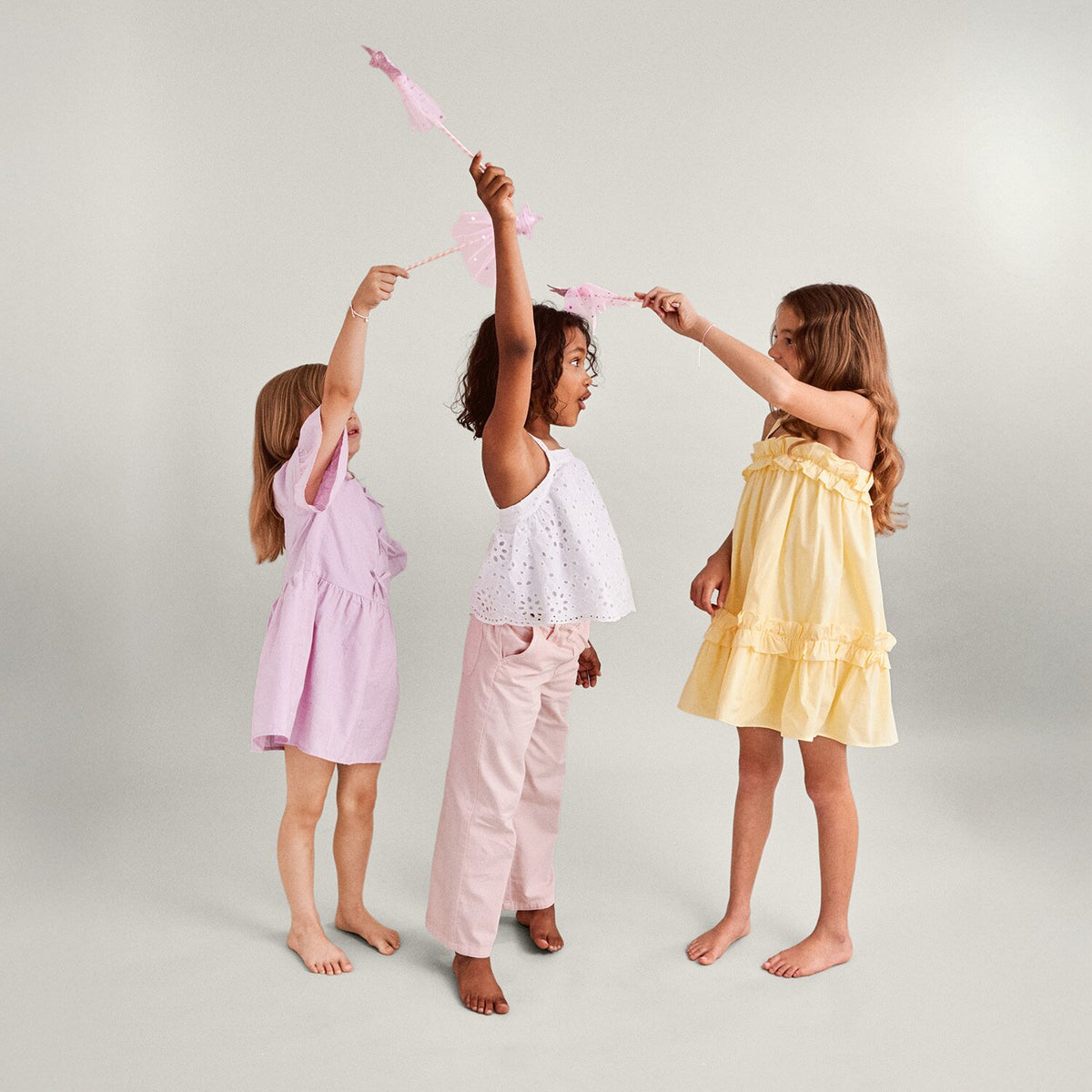 Three young girls in pastel dresses and pants holding pink fairy wands against a light background