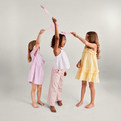 Three young girls in pastel dresses and pants holding pink fairy wands against a light background