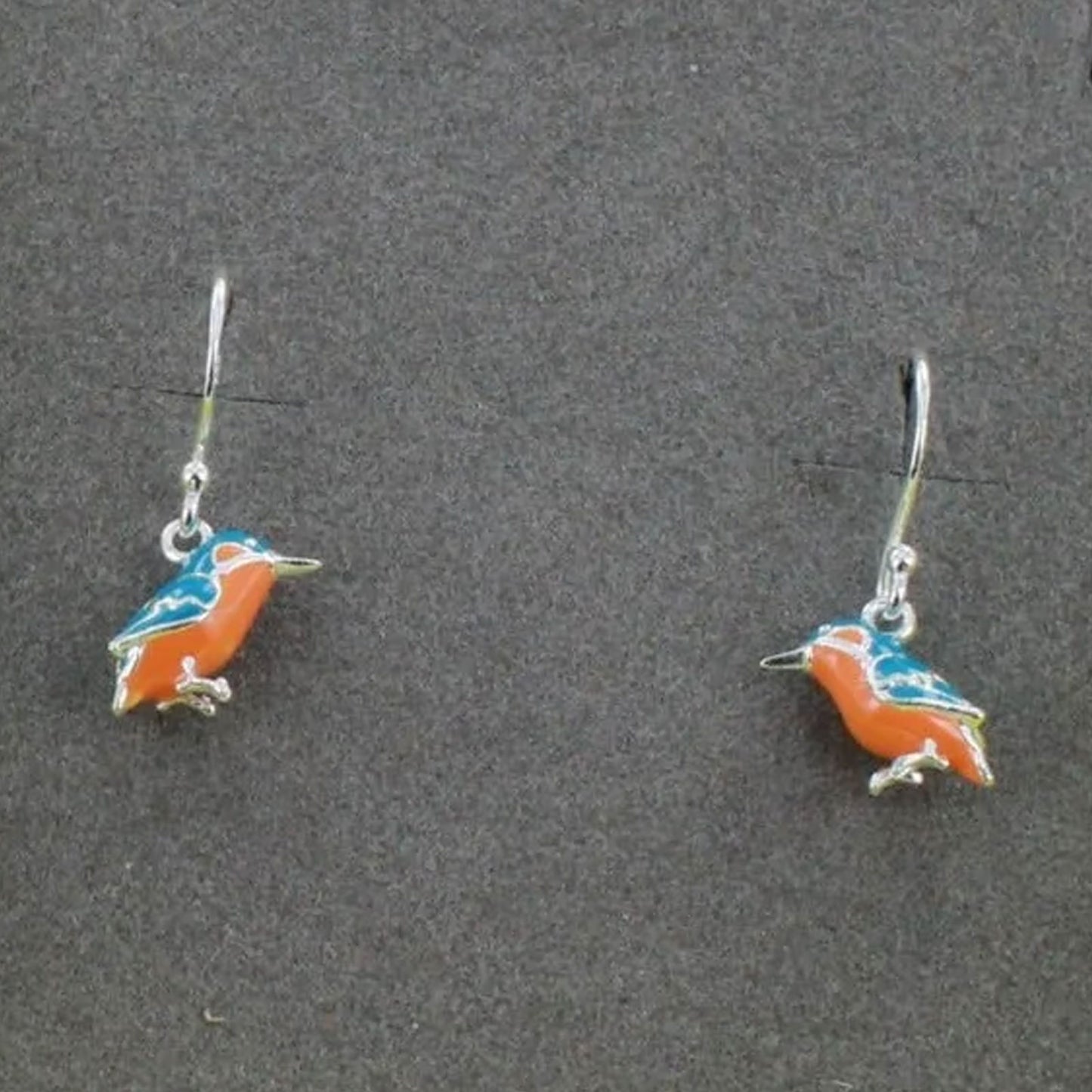 A pair of silver hook earrings featuring small bird charms with orange, blue, and white enamel details, displayed on a dark gray textured background.