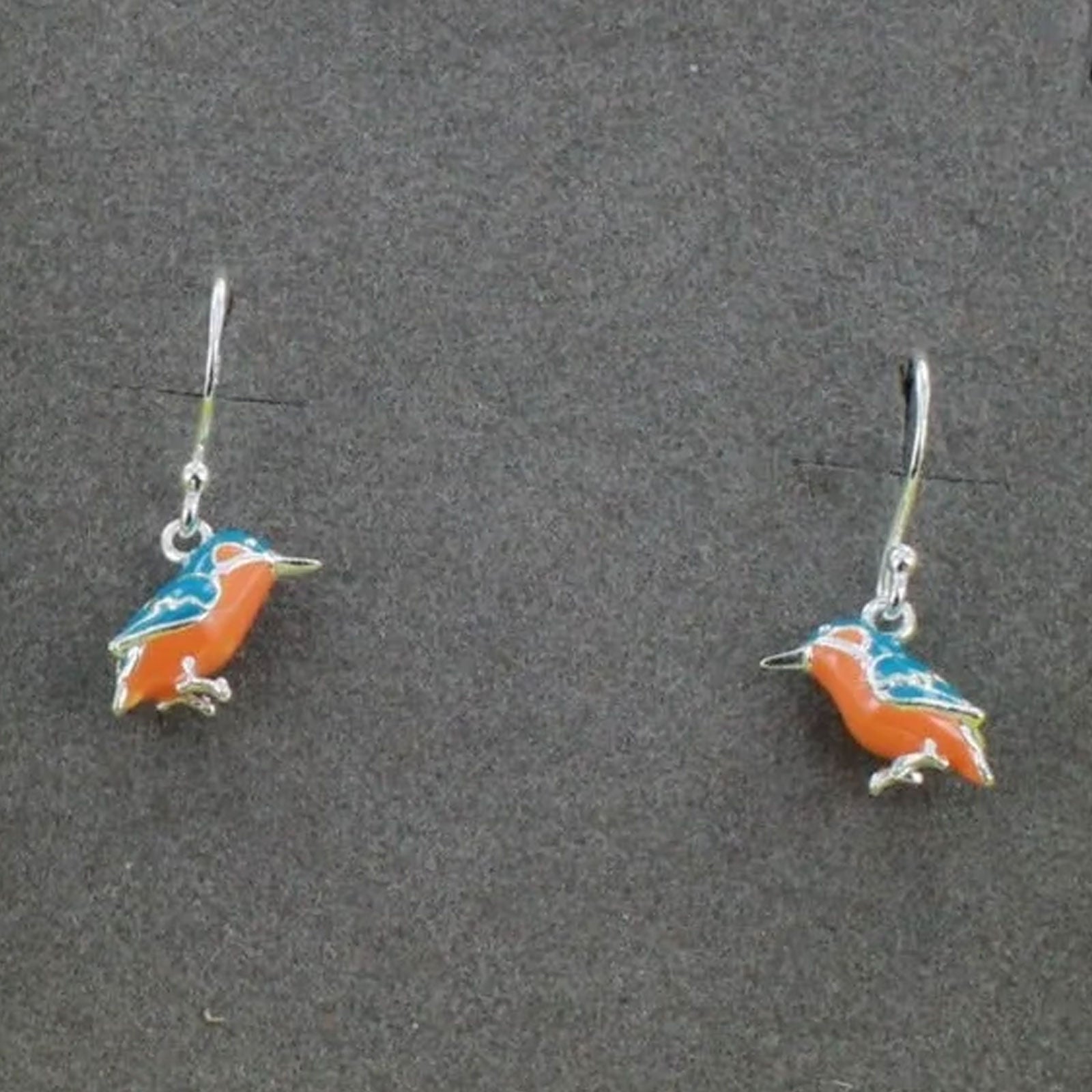 A pair of silver hook earrings featuring small bird charms with orange, blue, and white enamel details, displayed on a dark gray textured background.
