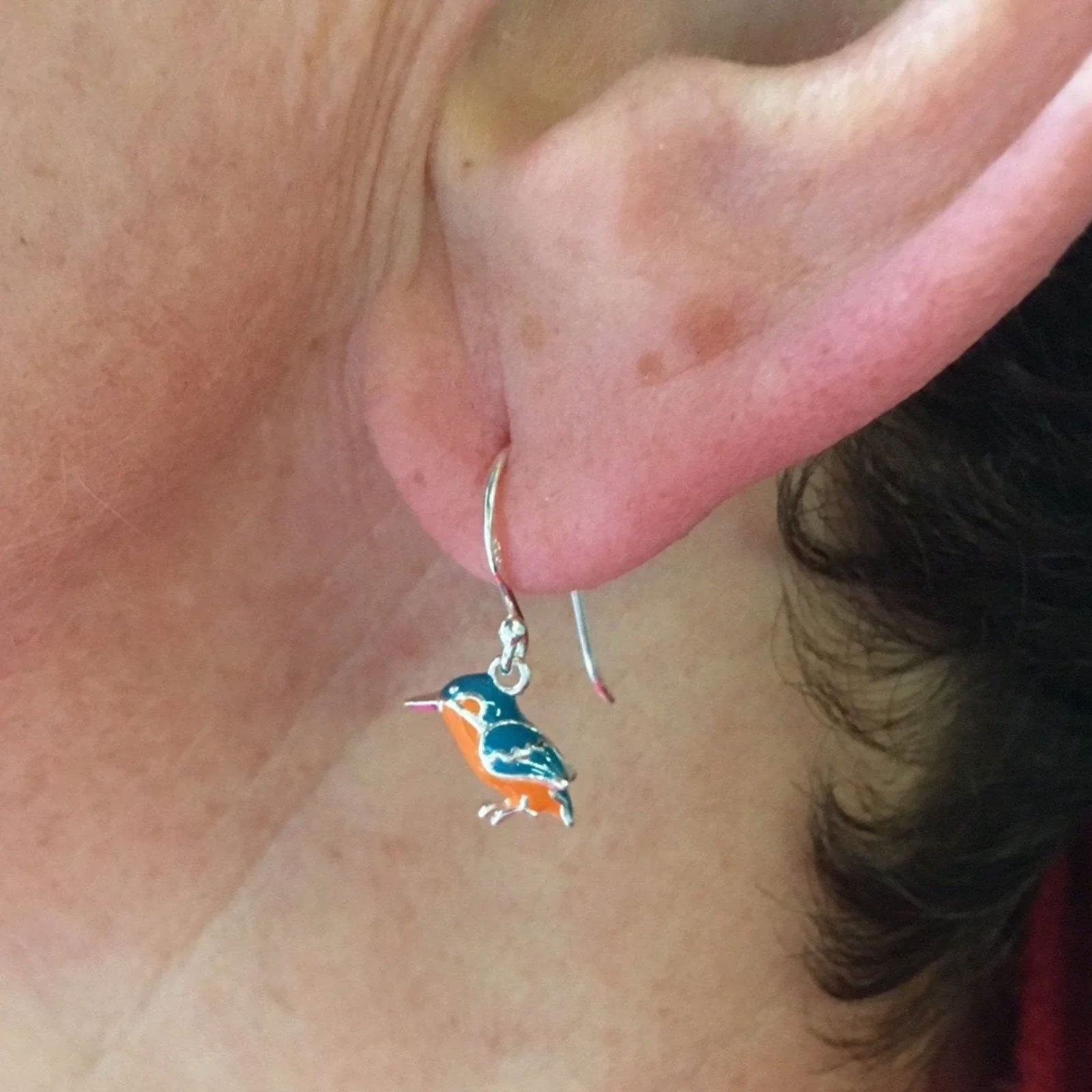 A close-up of a person's ear wearing a silver hoop earring with a dangling bird charm in blue, orange, and white enamel.