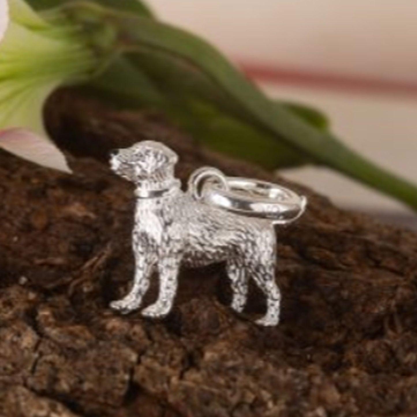 A silver dog-shaped charm or pendant with a textured finish and a smooth ring loop, displayed on a piece of bark with blurred green leaves in the background.