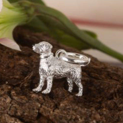 A silver dog-shaped charm or pendant with a textured finish and a smooth ring loop, displayed on a piece of bark with blurred green leaves in the background.