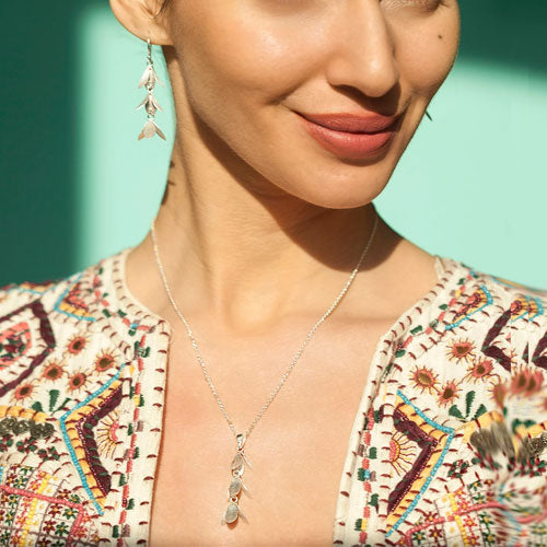 A close-up lifestyle shot of a woman wearing a delicate silver pendant necklace with three small shaped, matching dangling earrings, and a colourful embroidered top against a mint green background.