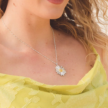 A close-up lifestyle shot of a woman wearing a delicate silver chain necklace with a daisy pendant. The pendant features a white center and silver petals, worn over a light yellow-green lace top.